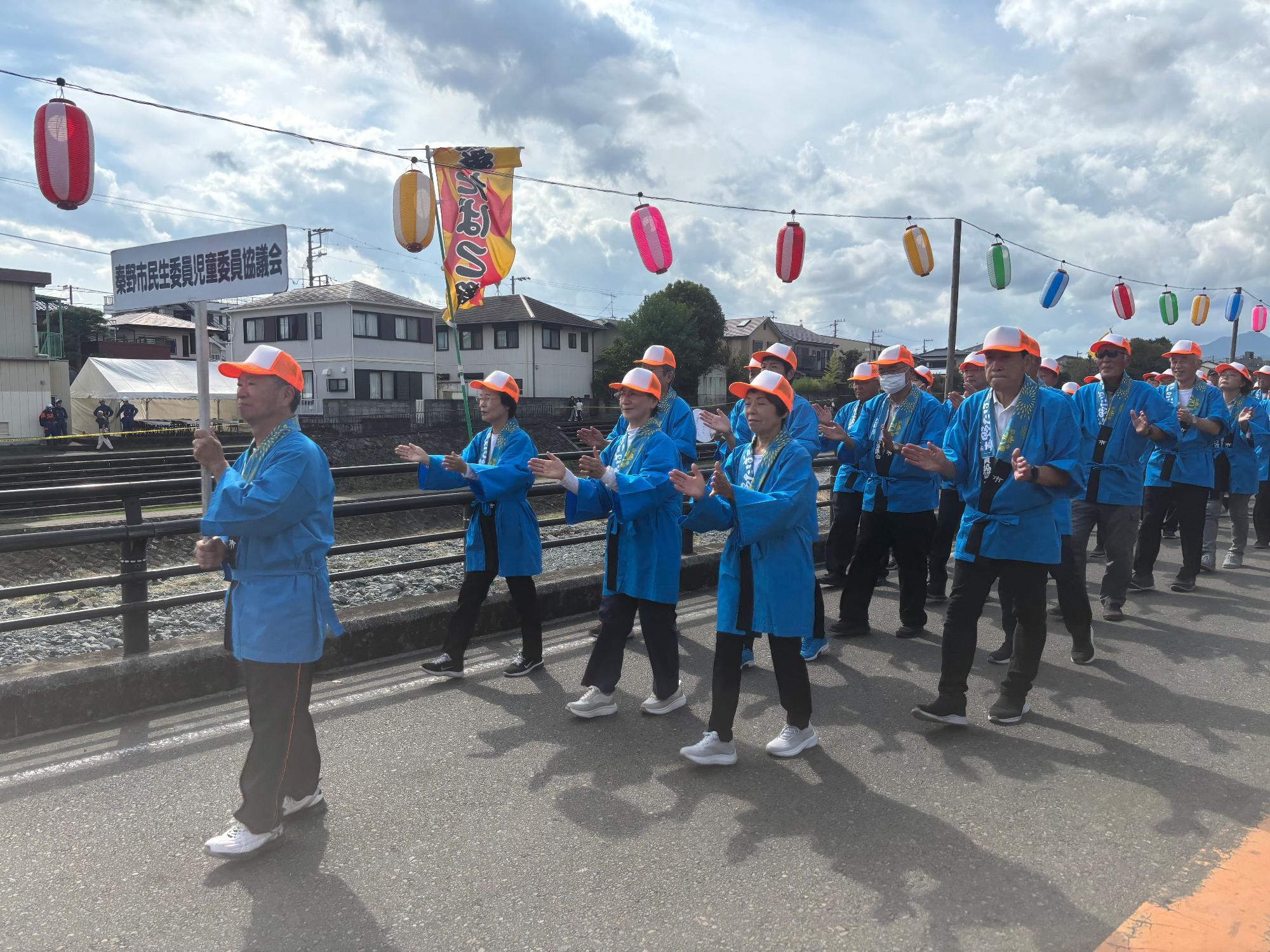 カラフルな提灯が飾られた道路で青い法被とオレンジ白の帽子を着た多数の踊り手が列をなして盆踊りを披露している写真