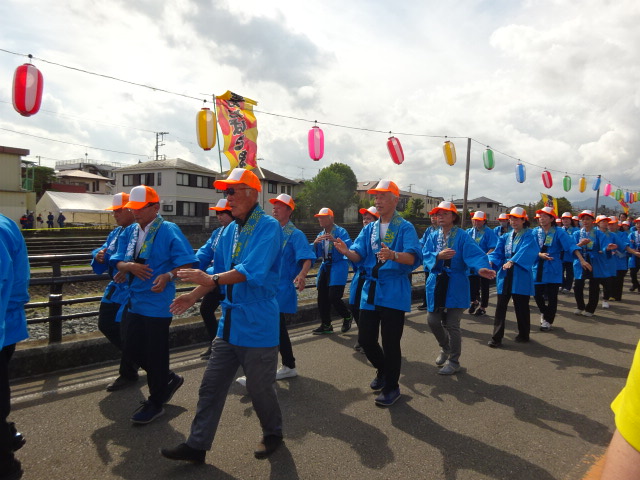 青い法被とオレンジの帽子を着用した人々が、カラフルな提灯が吊るされた通りを踊りながらパレードしており、曇り空の下で活気ある雰囲気が伝わる祭りの写真