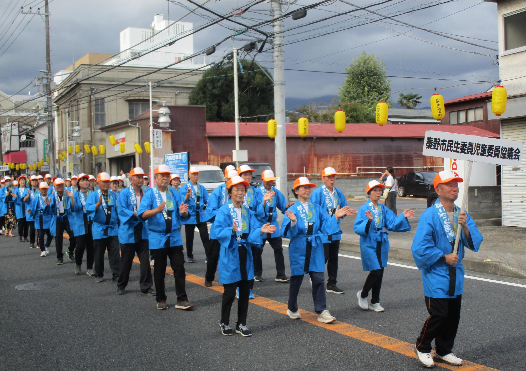 青い法被とオレンジの帽子を着用した人々が整然と隊列を組んで通りをパレードしており、背景には黄色い提灯と「秦野市民生委員児童委員協議会」と書かれたプラカードが見える、祭りの写真