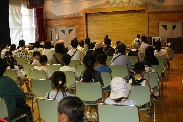 木製のステージがある広場で、大勢の子供と大人が淡い緑色の椅子に座って前方を見つめている風景を客席後方から捉えた写真