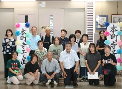 ドアの前に飾り付けされた「本町寺子屋」と書かれた看板が見える室内のロビーで、数名の高齢者を含む16人の男女が集合写真に収まっている集合写真