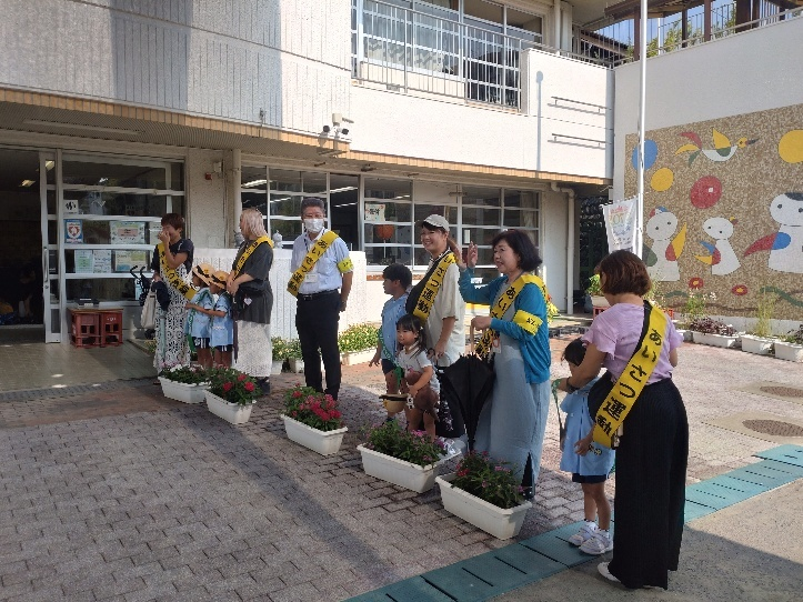 晴れた日の朝、幼稚園の玄関前で、子供たち数名と、「あいさつ運動」などのタスキをかけた大人のボランティアの人々が、植木鉢の前に横一列に並んで立っている挨拶運動の様子の写真