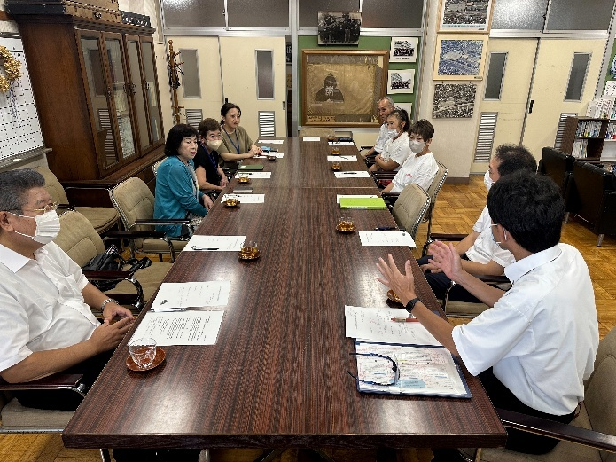 レトロな応接室のような場所で、資料や飲み物が置かれた長い木製テーブルを囲んだマスクを着用した男女9人が、真剣な表情で話し合っている会議の様子の写真