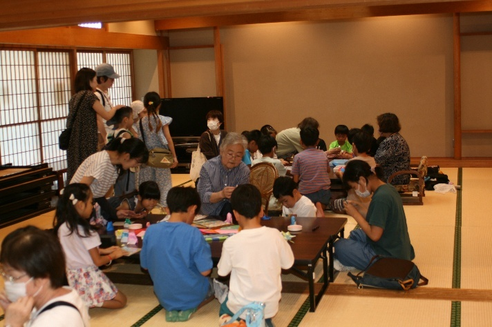 座卓が並んだ和室で、畳の上に大勢の子供たちと数名の大人たちが座り、折り紙などの活動を通じて地域交流を楽しんでいる様子の写真