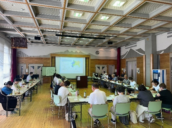 会議室の様な室内で、大勢の参加者が長机をコの字型に囲んで座り、前方のスクリーンに映し出された資料を見ている防災研修会の様子の写真