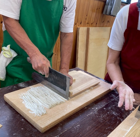 緑のエプロンをつけた男性が、木製のまな板に置かれた打ち立ての蕎麦を、蕎麦切り包丁を使って均一な細さで切っている、蕎麦打ち体験の様子の写真