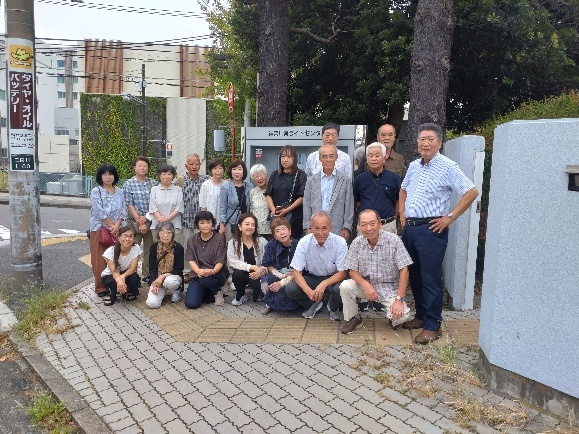 周囲に樹木が立ち並んだ神奈川県ライトセンターの入り口の前の歩道脇で、大勢の参加者が立ったりしゃがんだりして並んでいる集合写真