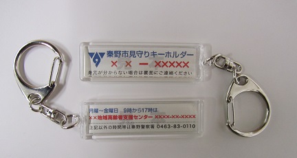 秦野市見守りキーホルダーと書かれた透明なプラスチック製ケースと銀色の金具からなるキーホルダーが、二つ並べて配置された写真