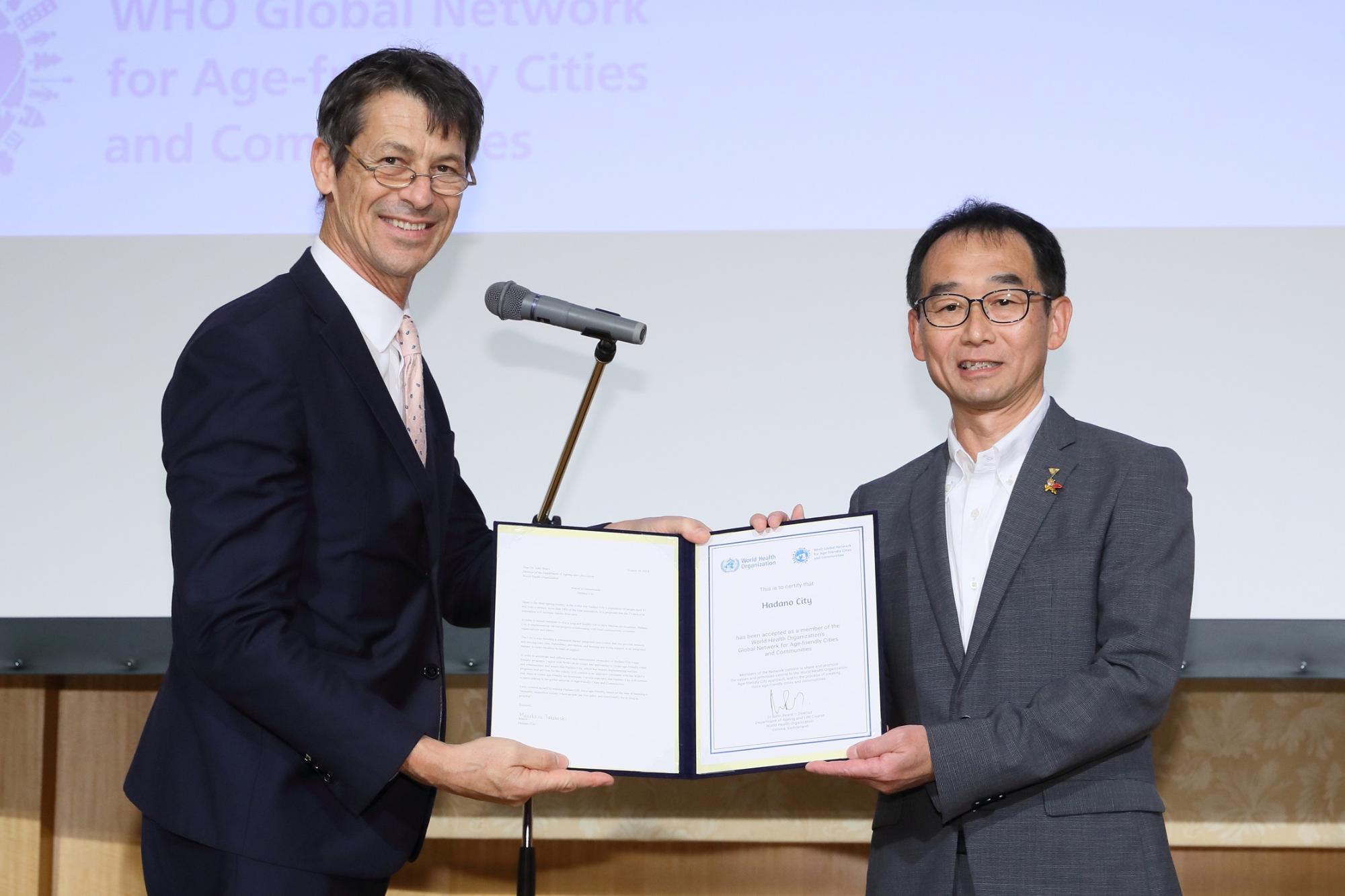 ジョン・ベアードWHOエイジング・アンド・ライフコース部長と、宮村副市長が並び、「エイジフレンドリーシティ参加承認証明書」を二人で持ち、笑顔でカメラを見ている様子の写真