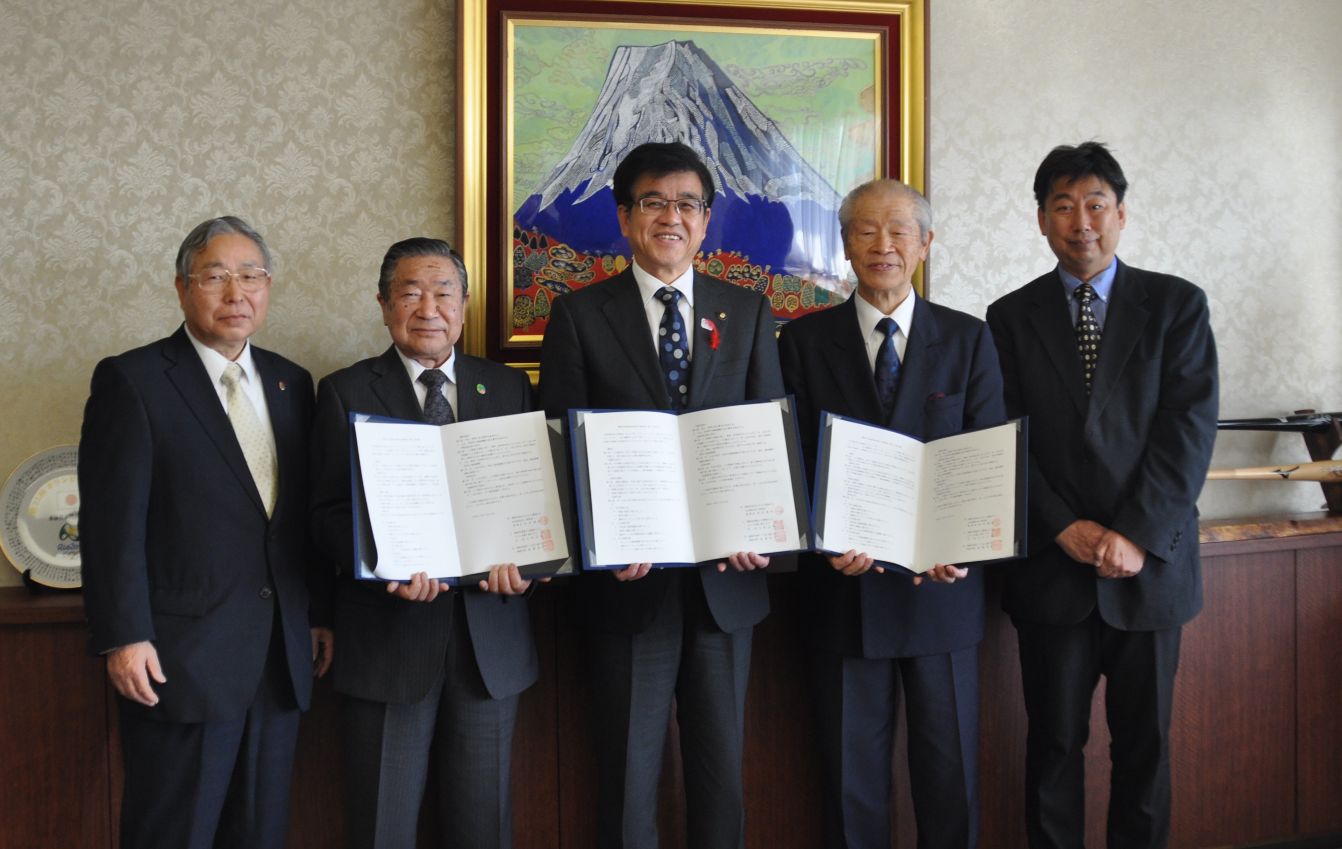 全員スーツ姿の5人の男性が中央の3人がそれぞれ開いた書類を持って並び、友好協定か覚書のような文書を締結した際の記念撮影と思われる室内の集合写真