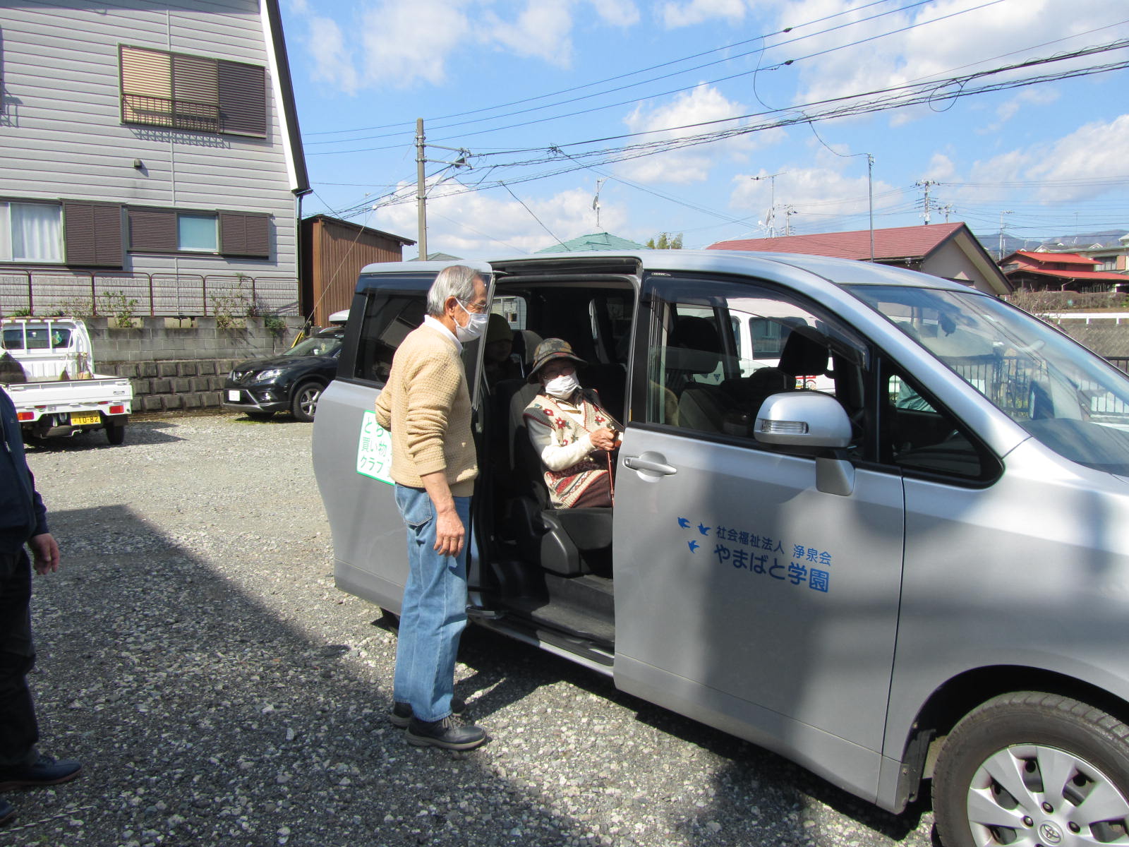 砂利の駐車場で、マスクを着用した男性が後方のスライドドアを開けて座っている高齢女性に声をかけている様子の写真
