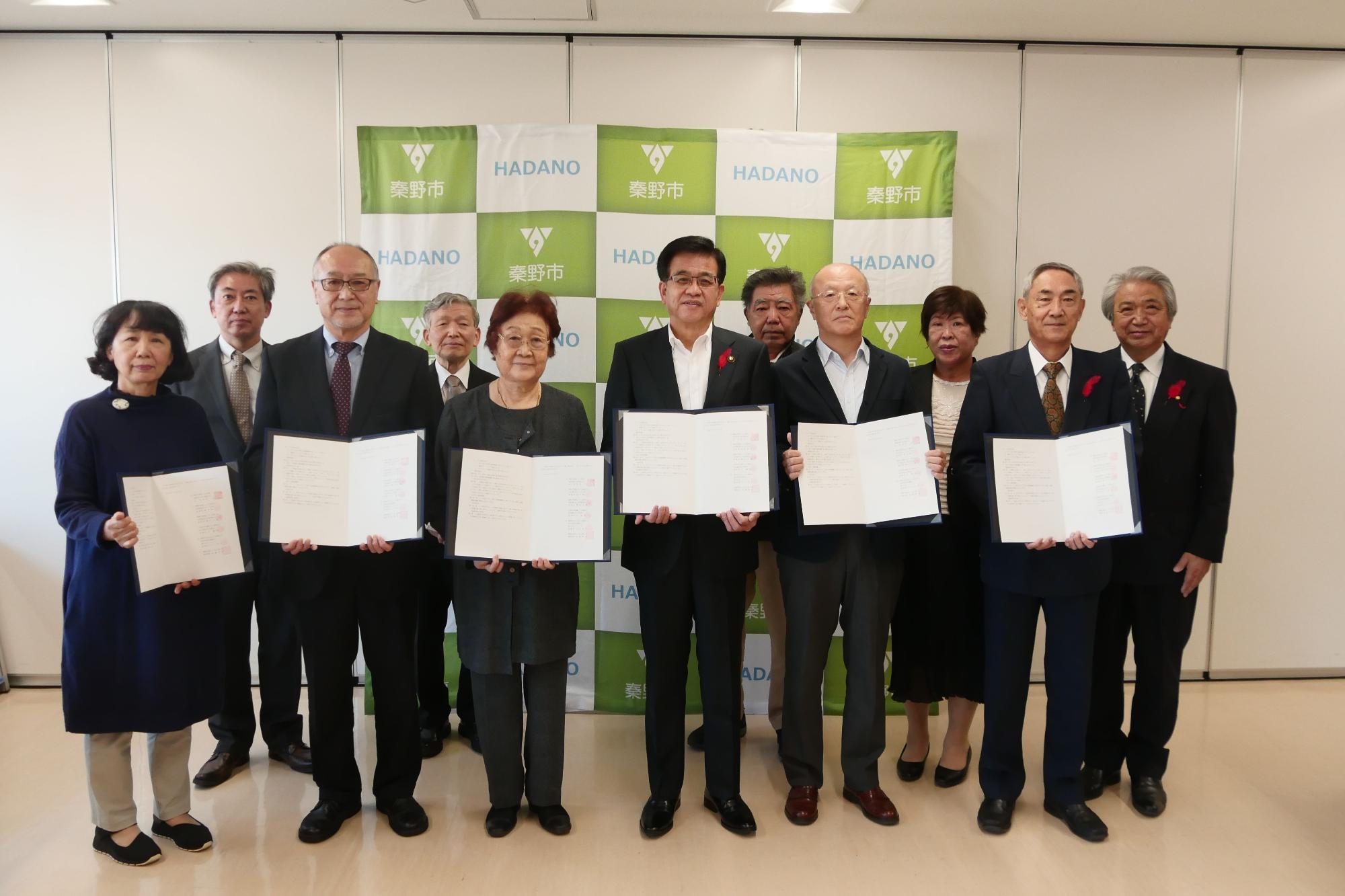 緑のグラデーションの秦野市（HADANO）ロゴが入ったバックパネルの前で、男女計11名が中央の4名が開いた文書を手に持ち、何らかの協定または覚書の締結式における記念撮影をしている集合写真