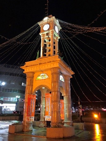 夜間照明と周囲に張り巡らされたイルミネーションが施された、花崗岩のような石材で造られた四面時計台の写真であり、土台部分にはオレンジ色ののぼり旗が立てられ、背景には街の建物や明るい光が見えるランドマークの写真