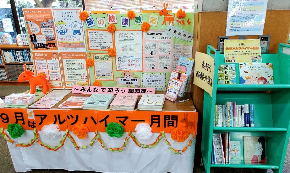図書館のような室内に設けられた、9月は世界アルツハイマー月間というテーマの啓発展示の全体像を捉えた写真であり、オレンジと白と緑の装飾が施されたテーブル上には認知症に関するパンフレットや書籍が並び、背景の掲示物には「脳の健康教室」の案内や卒業生の声などが書かれている展示風景の写真