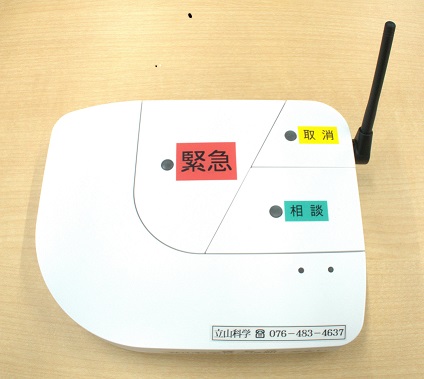 赤色の「緊急」、黄色の「取消」、緑色の「相談」のラベルが貼られたボタン配置部分と、右上に飛び出した黒いアンテナがある緊急通知装置本体の上面写真