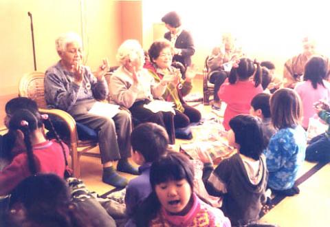 畳敷きの室内で、和やかな雰囲気の中、数人の高齢者が椅子に座って拍手をし、その周りの床には座ったり寝転んだりしている大勢の子どもたちが楽しそうに集まっている写真