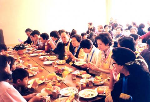 長くて茶色いテーブルを囲むように、中央に高齢者が集まり、手前には数人の子どもたちも混じって座っており、皆が笑顔で食事を楽しんでいる写真