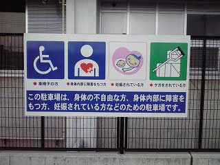 建物前のフェンスに設置された、四角で囲まれた、車いすマーク、ハートプラスマーク、マタニティマーク、けがをしているひとのマークが横並びに掲載され、下に「この駐車場は、身体の不自由な方、身体内部に障害をもつ方、妊娠されている方などのための駐車場です。」と書かれた看板の写真
