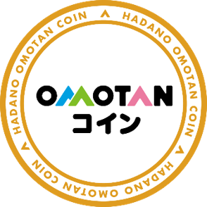 茶色い円形の縁に「HADANO OMOTAN COIN」という文字が書かれ、その内側の白い円の中央に「OMOTAN」が青、緑、ピンク、黒の文字色で、「コイン」が黒文字で書かれた秦野市デジタル地域通貨のロゴマークの画像