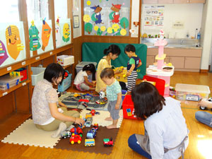 室内のプレイルームのような場所で、畳敷きの床に座った数名の大人と子供たちが、おもちゃの車やブロックなどで遊んで交流しており、壁には明るい装飾が施されている写真