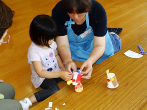 木の床の上で、一人の女性が子供と一緒に、紙コップや色紙などを使って工作を楽しんでおり、女性が手を添えて子供の製作を手伝っている様子の写真