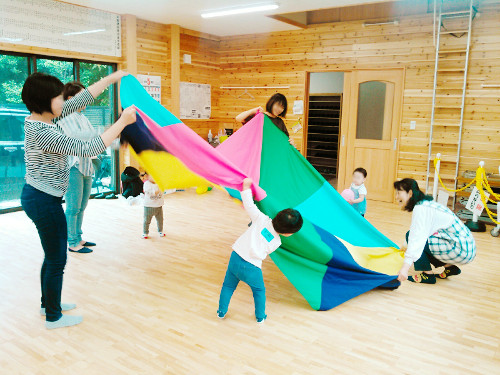 広くて明るい木目調の部屋で、数人の大人と幼児たちが集まり、カラフルな布を使ってパラシュート遊びのようなレクリエーションを楽しんでいる様子の写真