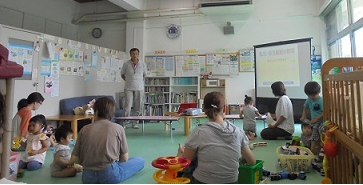 床に座る親子の周りに様々なおもちゃや育児用品が置かれた広々としたプレイルームで、立っている男性が壁のスクリーンに資料を投影しながら、育児中の親たちにミニミニ講座を開いている様子の写真