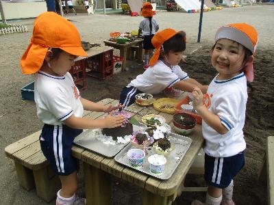 オレンジ色の帽子をかぶった4歳児が、テーブルに砂で作ったケーキやデザートを並べてお花で飾り付けして遊んでいる写真