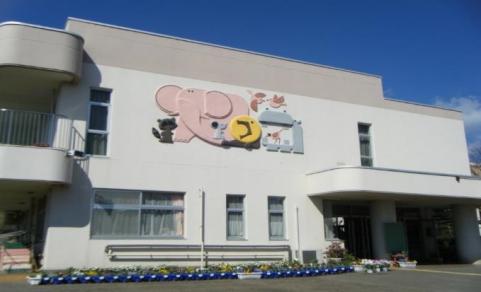 明るい青空の下、白い壁の二階建ての園舎の正面に、ピンクの大きな象やネズミ、狸やカバなどの動物の立体的なイラストが飾られている、親しみやすいデザインのつるまきこども園の園舎の建物を写した写真
