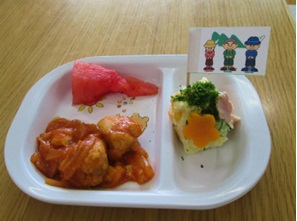 白いトレイに鶏肉のケチャップ煮・スイカ・丹沢はだの三兄弟が描かれた旗が立っている丹沢ポテト山盛りサラダが盛り付けられた、山の日の乳児のメニューの給食の写真