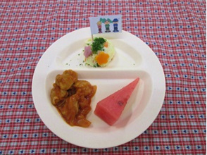 丸くて白いトレイに鶏肉のケチャップ煮・スイカ・丹沢はだの三兄弟が描かれた旗が立っている丹沢山盛りポテトサラダが盛り付けられた、山の日の幼児のメニューの給食の写真