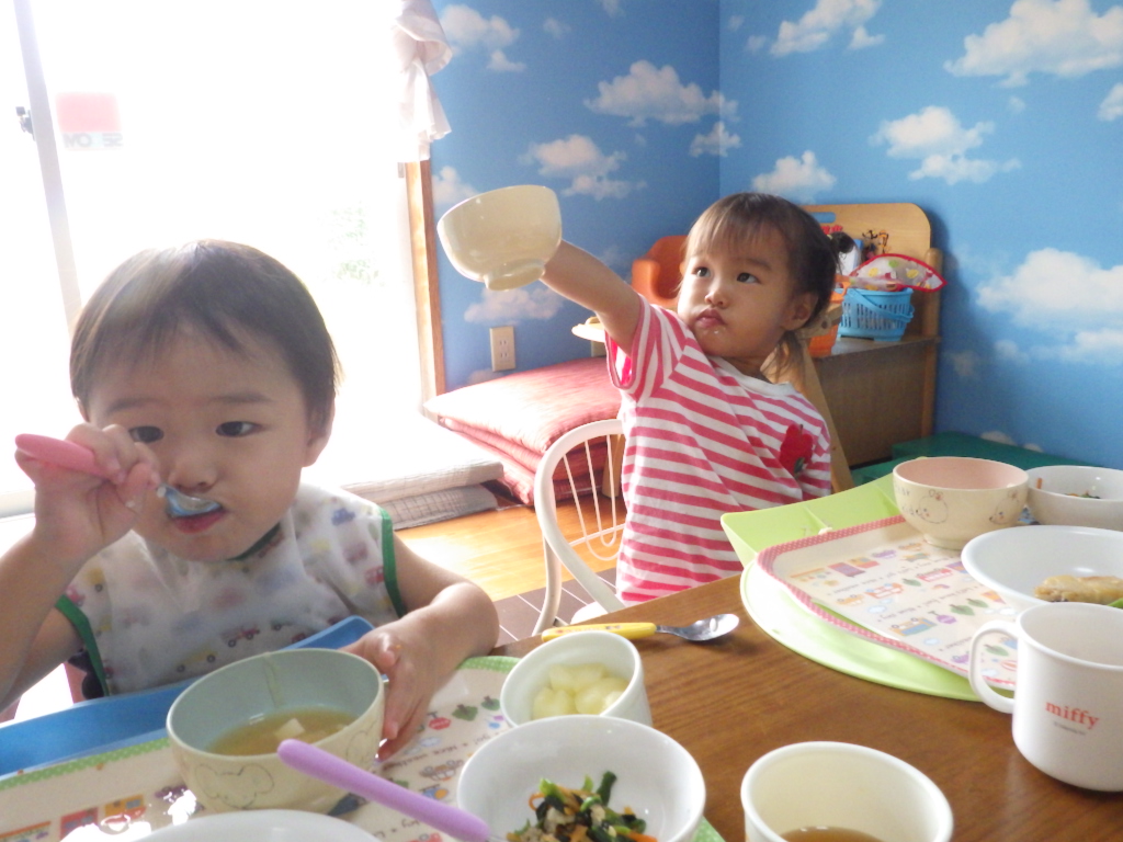青い空と雲の壁紙の部屋で、二人の幼児が木製のテーブルで食事をしており、手前の子供がスプーンでスープのようなものを食べている一方、奥の赤いボーダーの服の子供は片手を上げ、茶碗を差し出している、食事の様子の写真