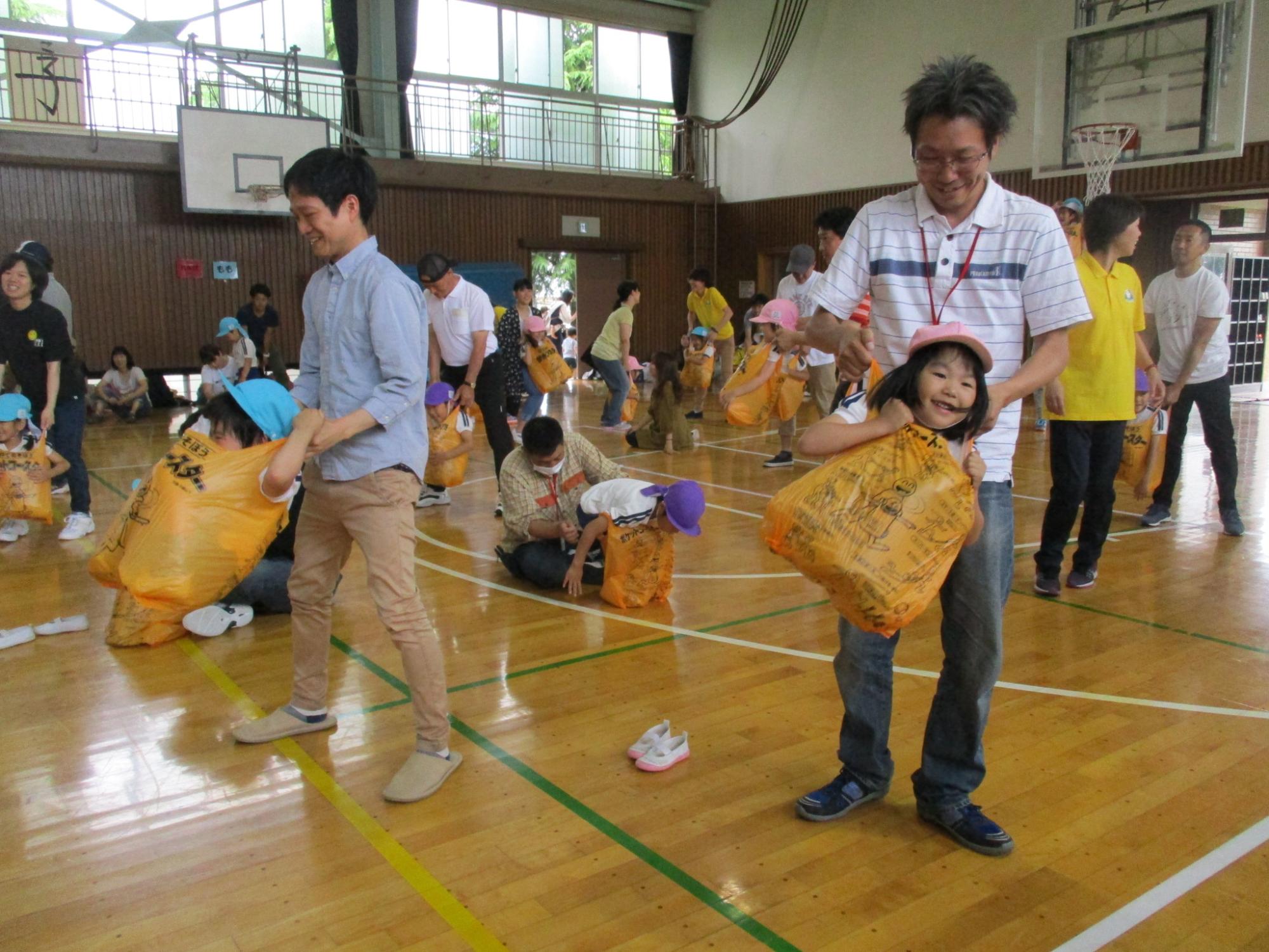 体育館で大勢の親子が大きなオレンジ色の袋に子供を入れて親が持ち上げ、楽しそうにレクリエーションに参加している写真