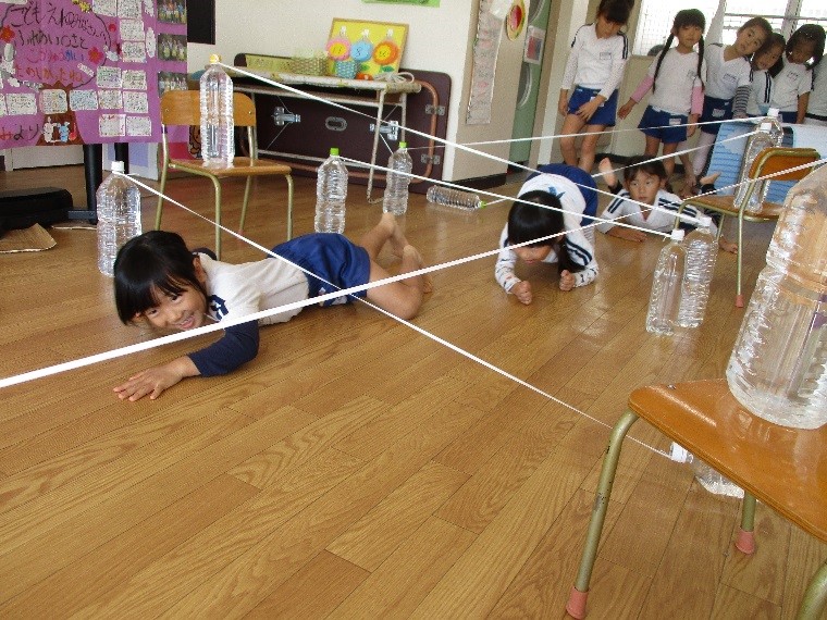 複数の子供が部屋に張り巡らされた白いひもを避けるように、床を這って楽しそうに遊んでいる写真