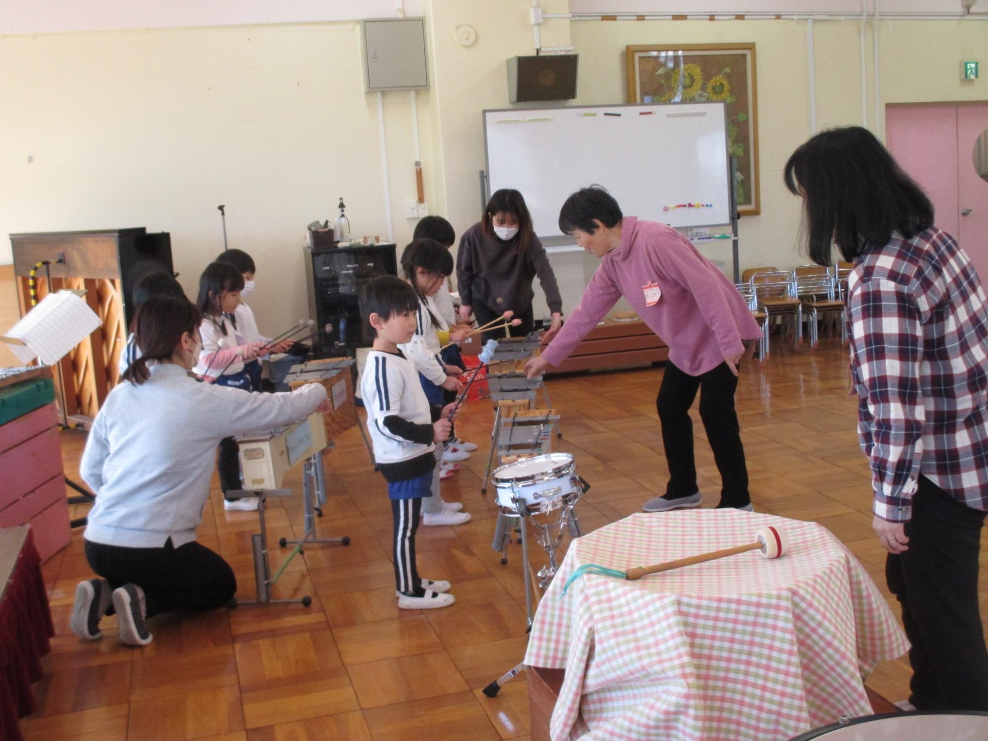 子供たちが木琴や太鼓などの楽器を使って、先生に指導されながら音楽を学んでいる教室での様子の写真
