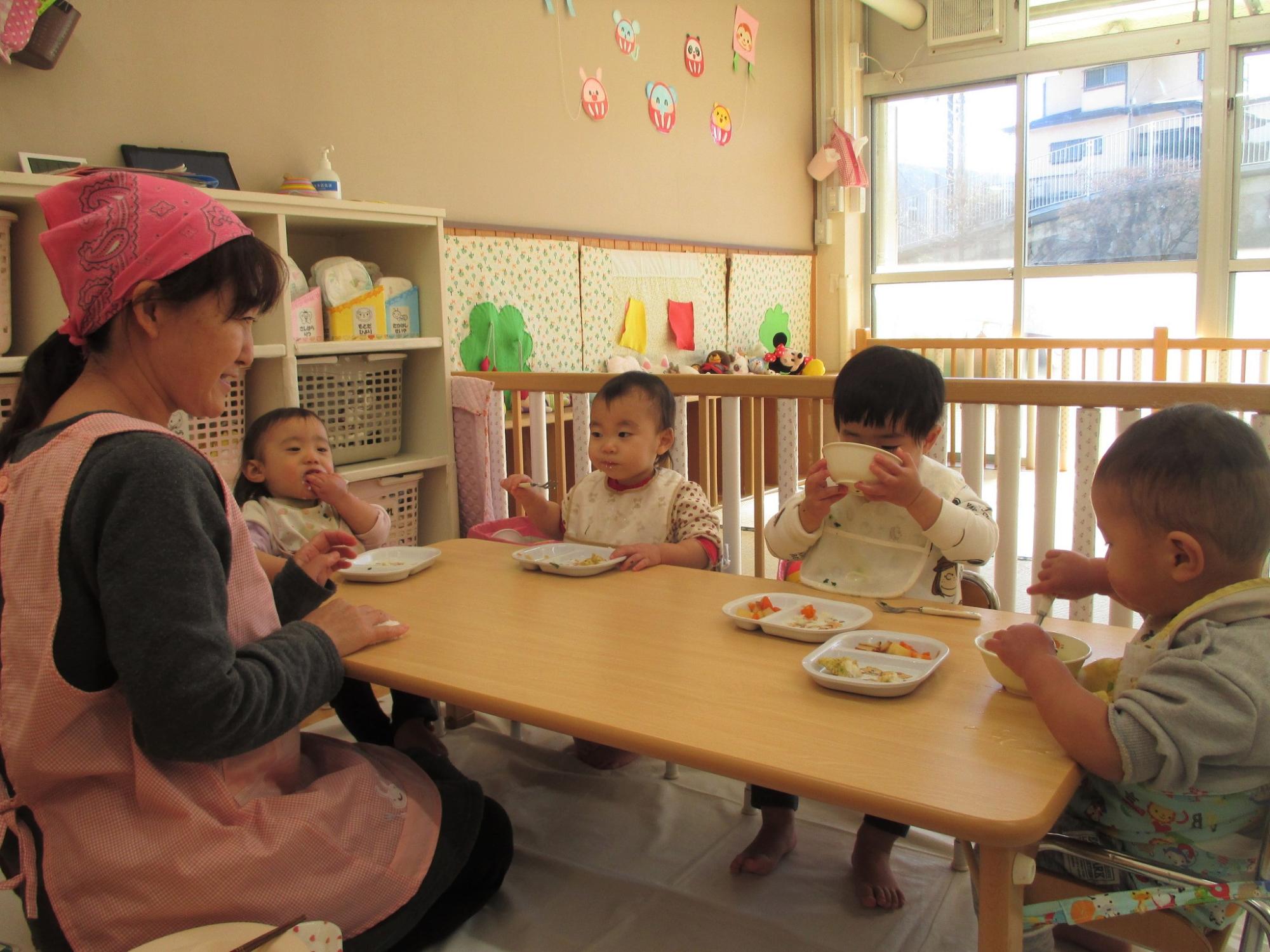 ピンクのエプロンとバンダナを身につけた保育士が、木製のテーブルで食事をしている複数の幼い子供たちを笑顔で見守っている写真