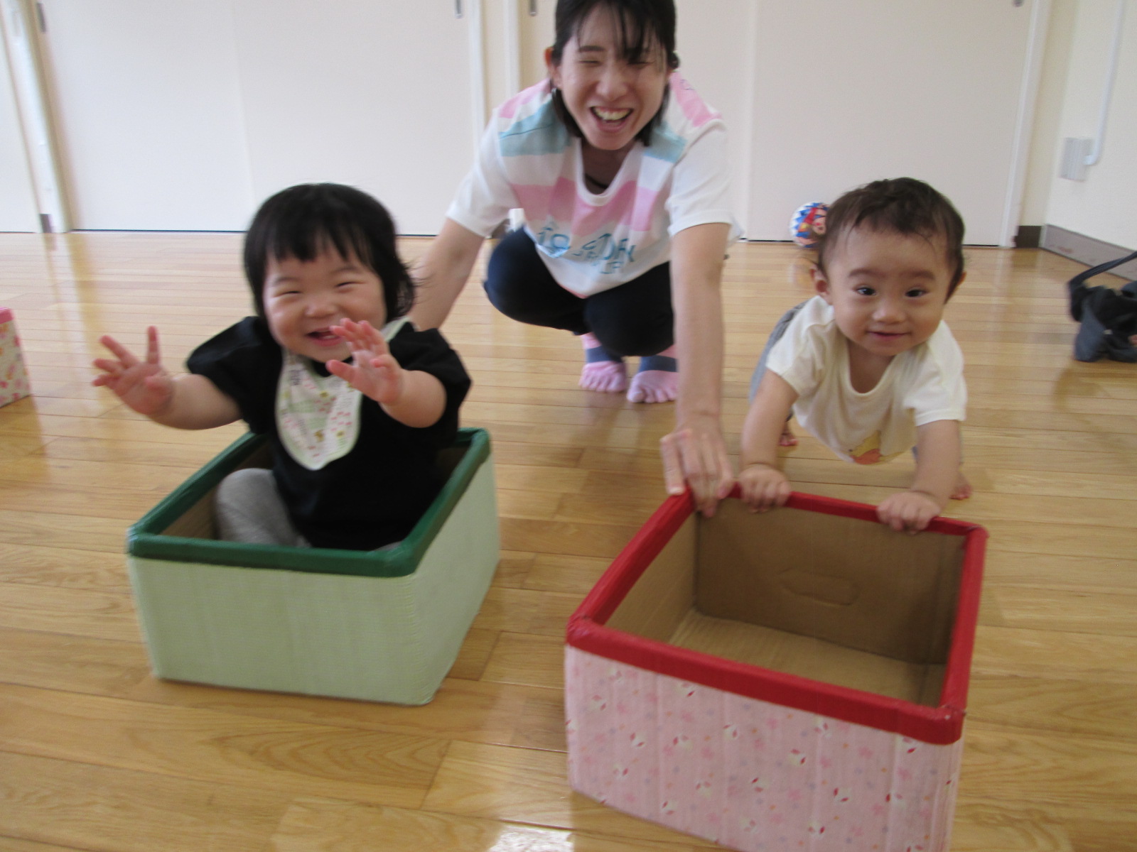 箱を乗り物に見立てて、楽しそうに笑いながら遊ぶ2人の幼い子供と、それを見守る保育士の先生を写した写真