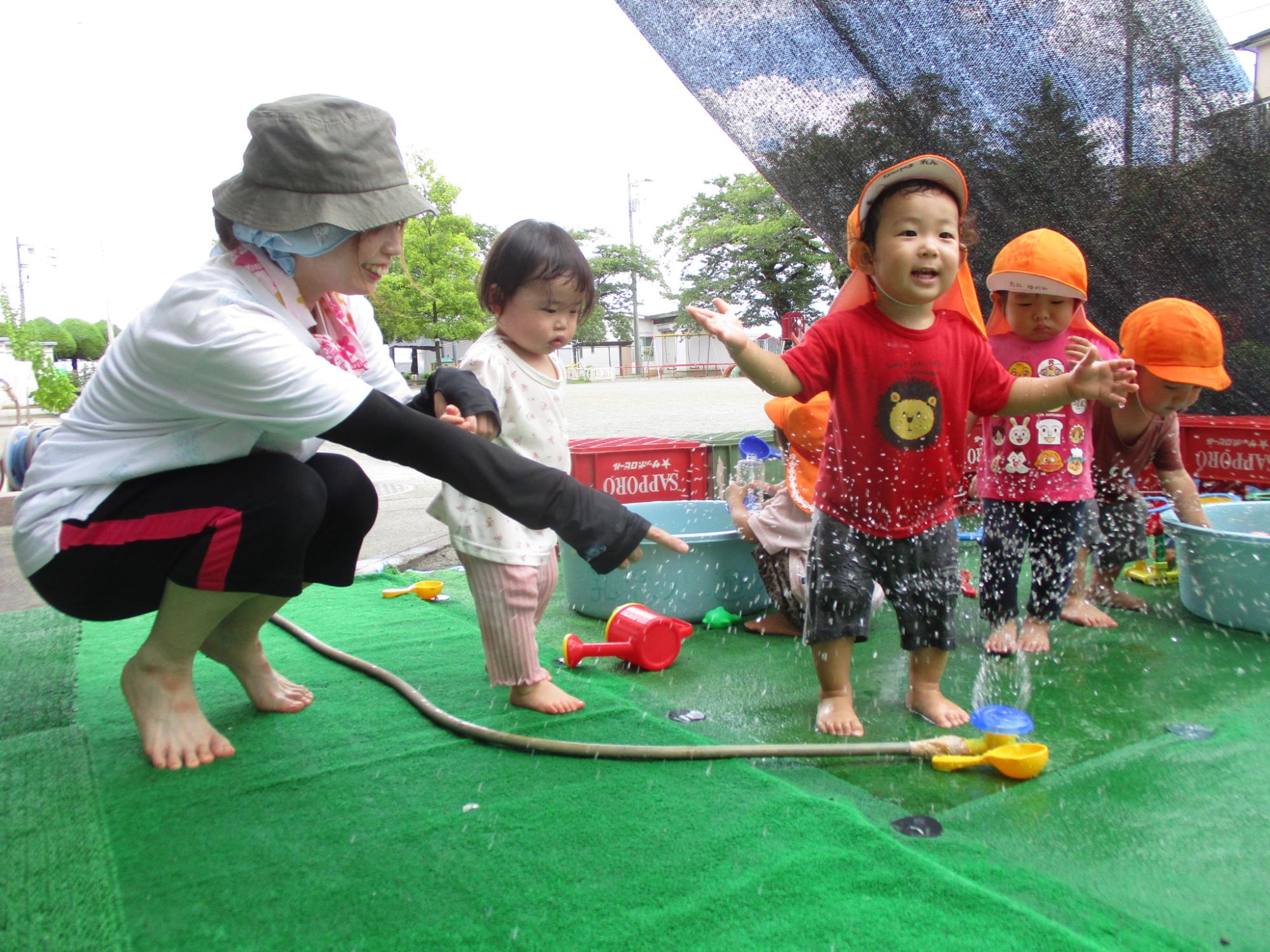先生に見守られながら、人工芝生の上で裸足の幼い子供たちが水遊び用のホースやバケツで楽しそうに遊んでいる写真