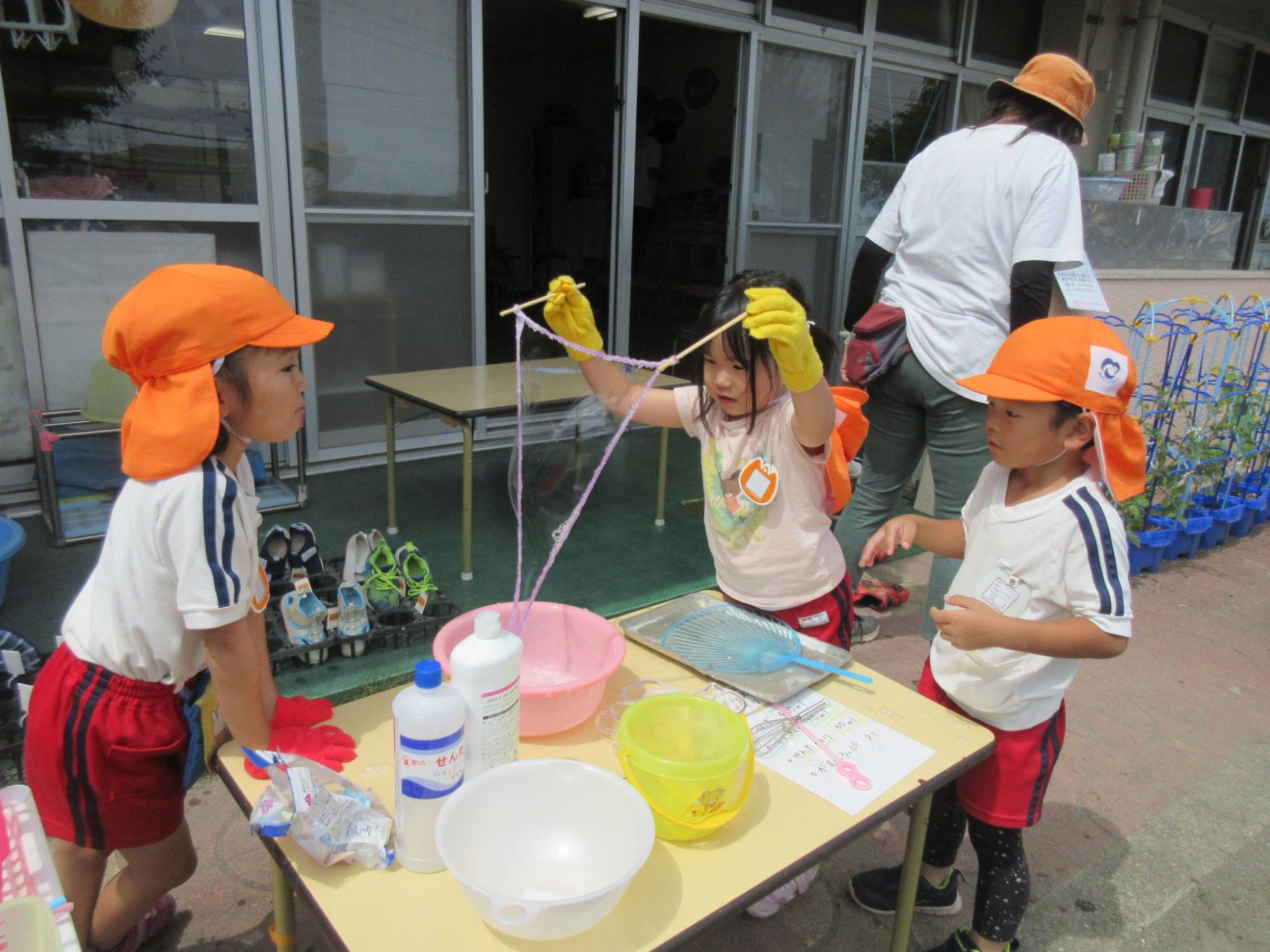 園庭に置かれた机でオレンジ色の帽子をかぶった3人の園児が、シャボン玉遊びをしている様子の写真