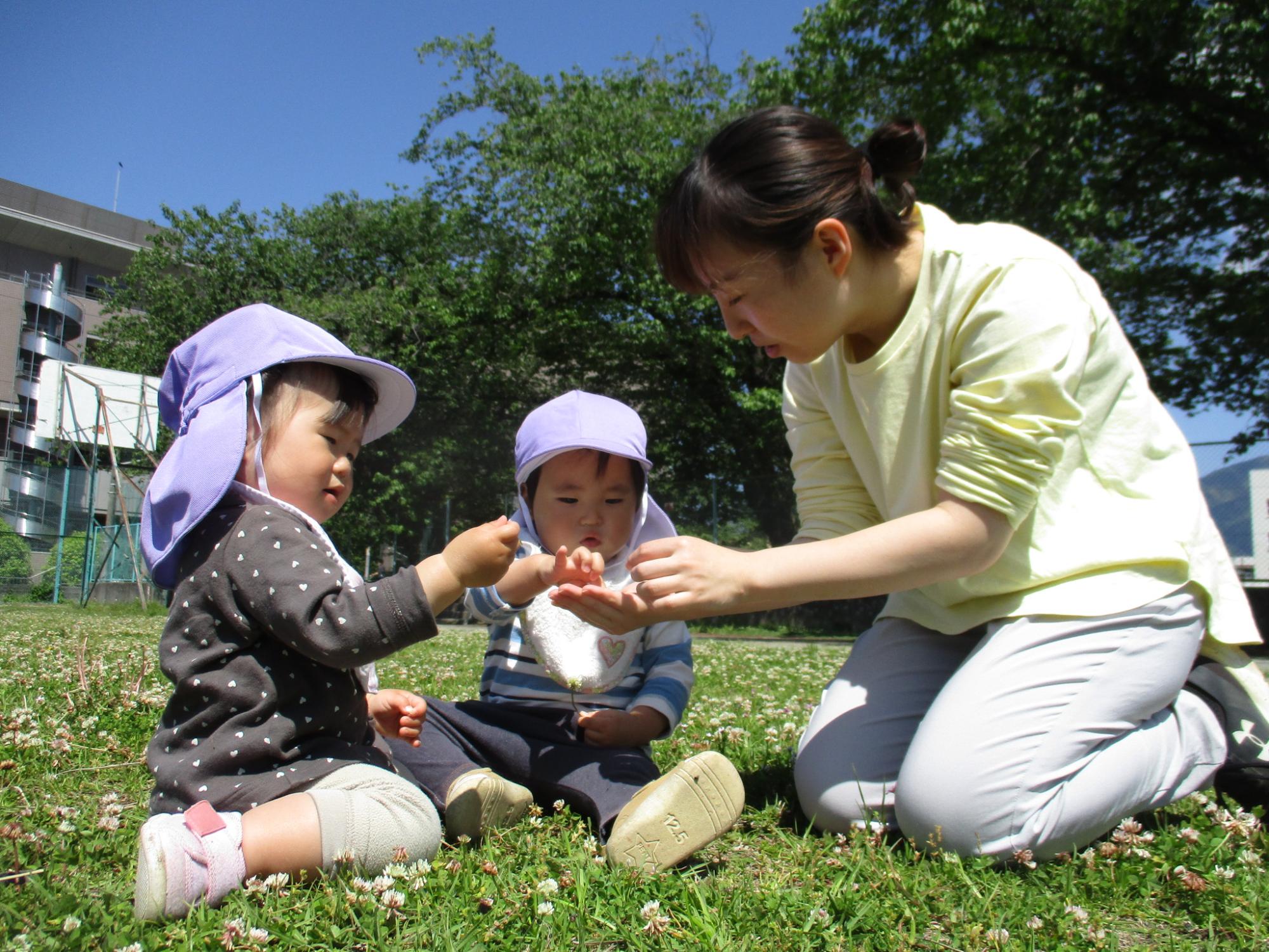緑の芝生に咲いた白い花に囲まれて、紫色の帽子をかぶった2人の園児が先生と楽しそうに遊んでいる様子の写真