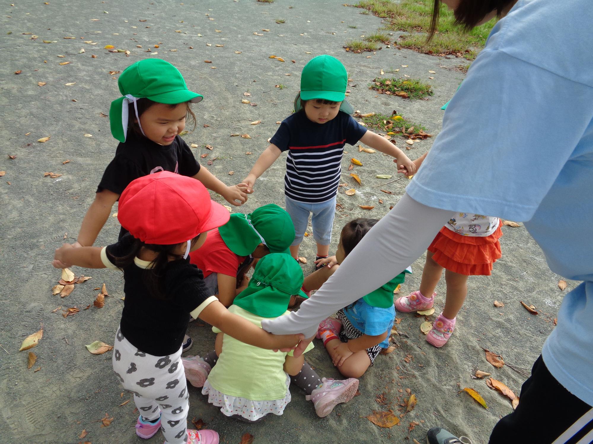 園庭で園児たちがしゃがんで座っている周りを他の園児たちと先生が、輪になって手をつなぎ囲んで遊んでいる様子の写真