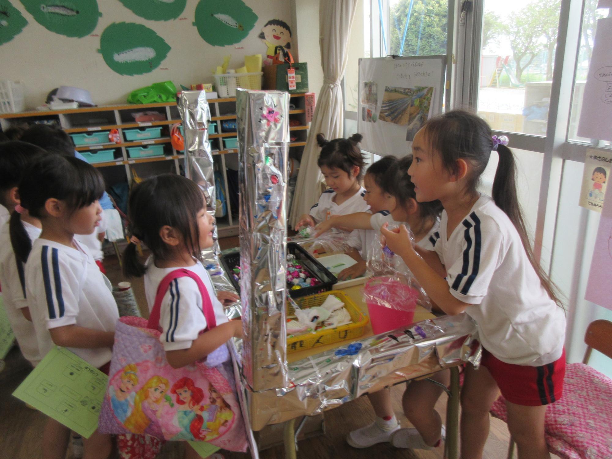 アルミホイルで装飾された工作物を作り、園児たちが店員とお客さんになり宝石屋さんごっこをして遊んでいる様子の写真