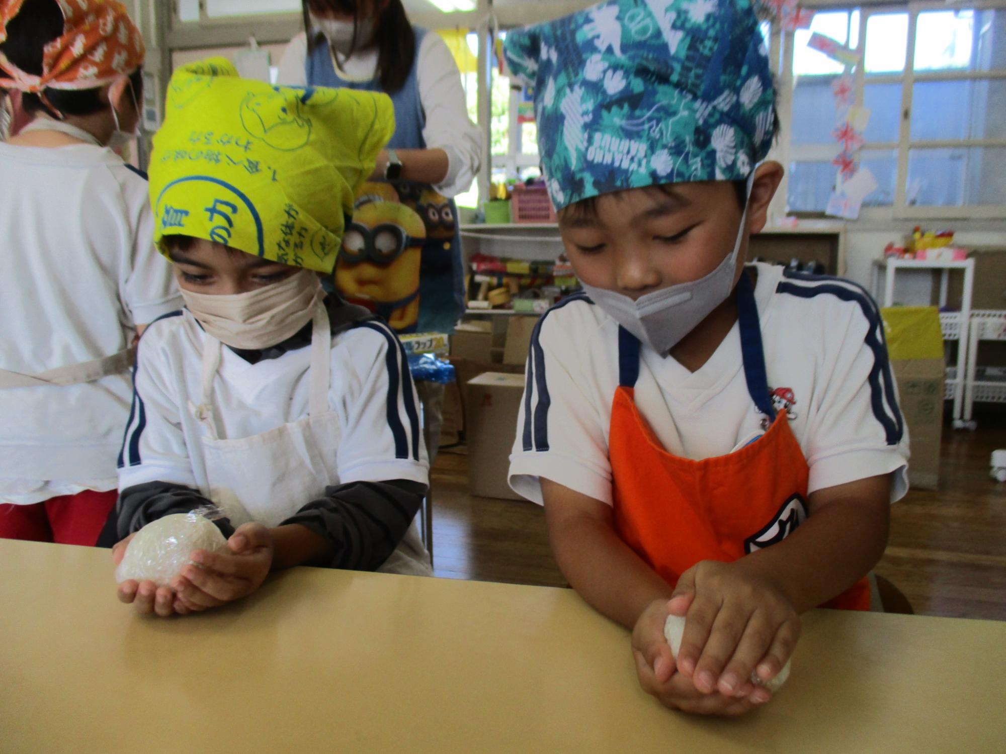 カラフルな三角巾とエプロンをつけた2人の園児が、机の上で白いご飯を握っておにぎりを作っている様子の写真