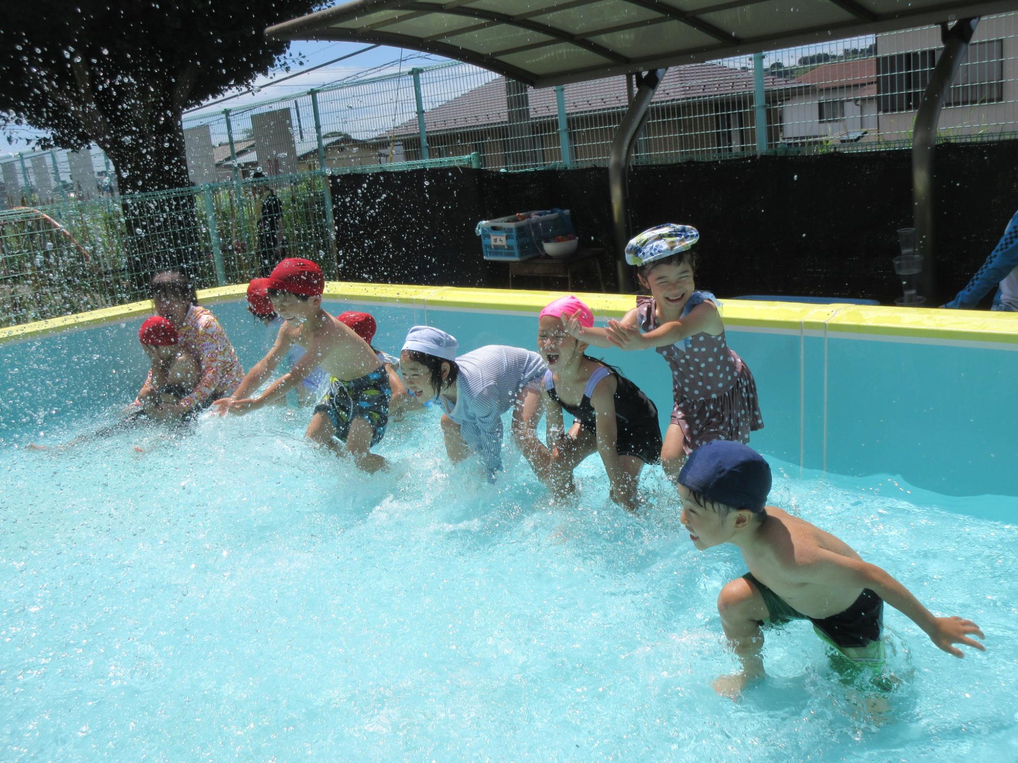 プールの中で、水着を着た園児が両手で水をすくい上げて水しぶきを上げながら遊んでいる様子の写真