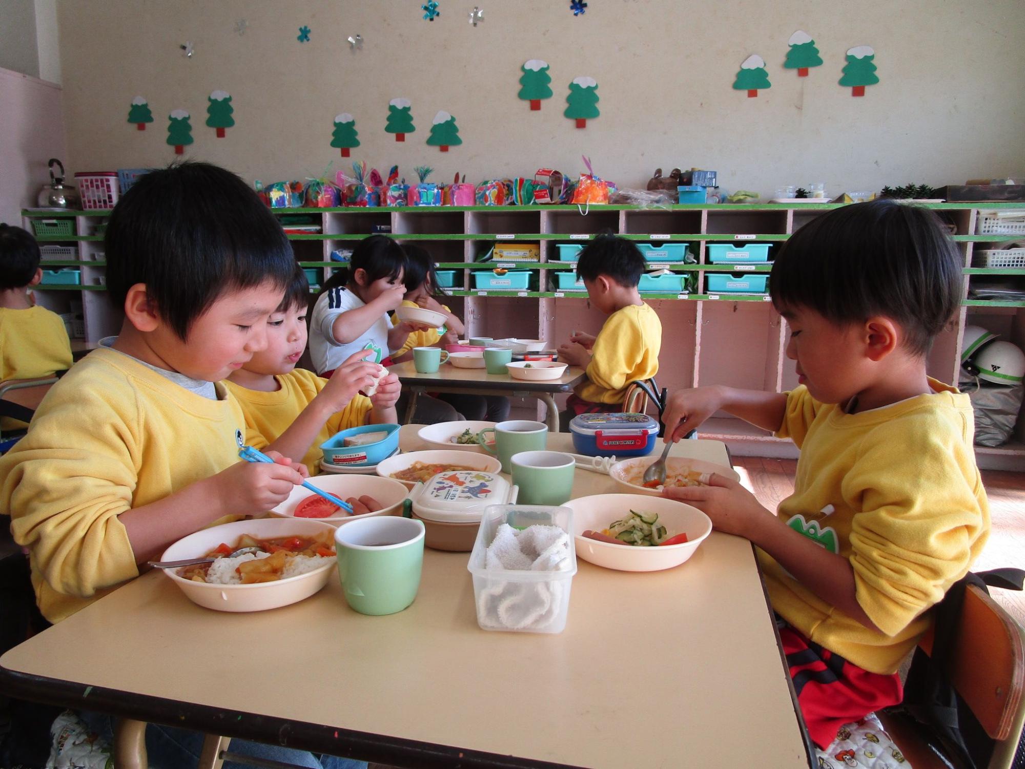 部屋の中で複数の園児たちが机に座り、それぞれスプーンやフォークを使って美味しそうに給食を食べている写真