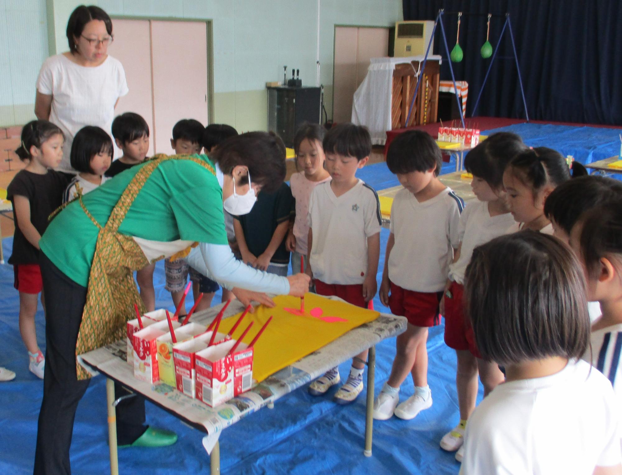 机の周りに集まった複数の園児たちに、講師が黄色い画用紙に絵を描いているのを見ている写真