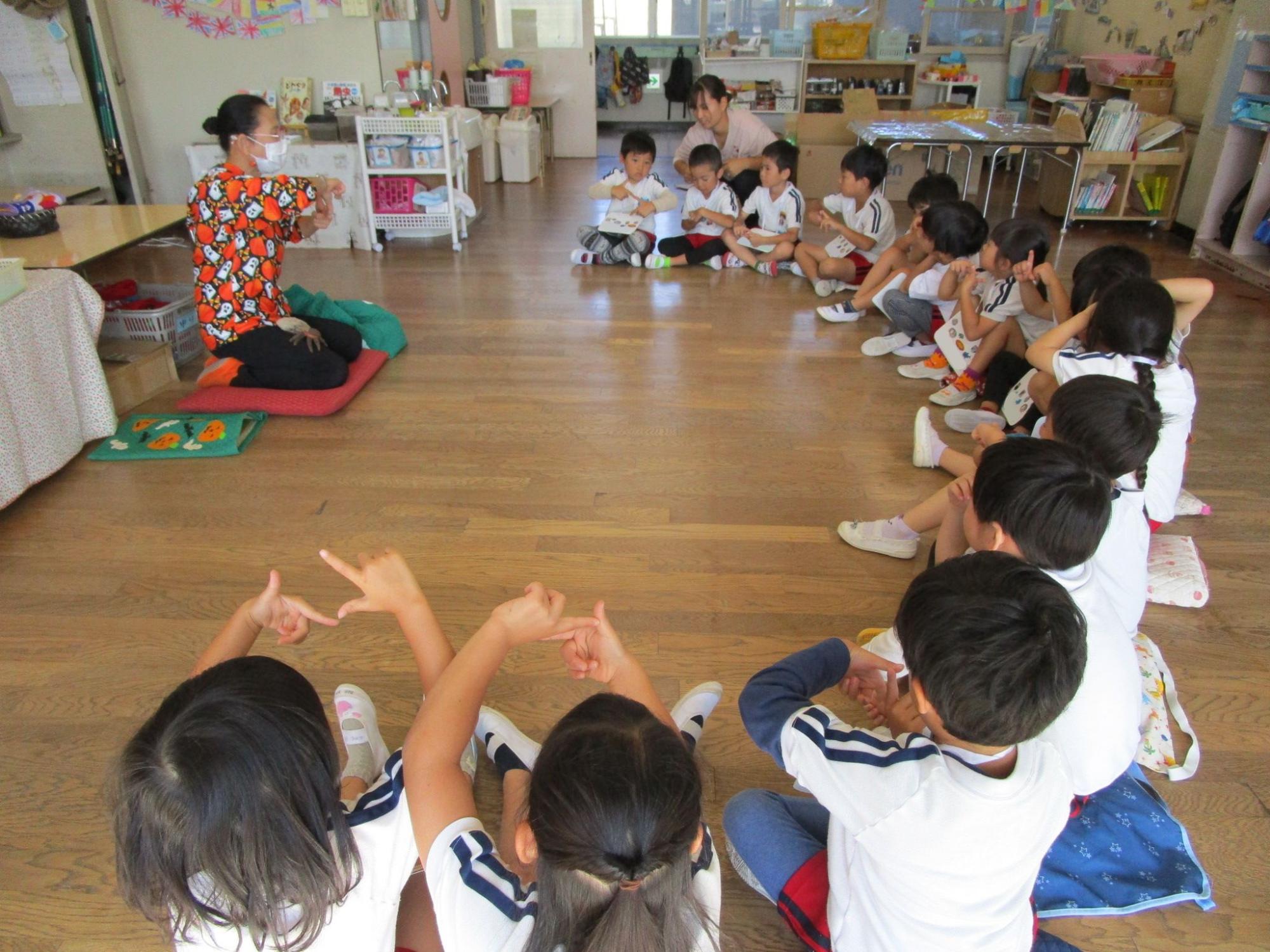中央の前に講師が座りそれを囲うように半円になって床に座った複数の園児が、講師の指導に合わせて楽しそうに両手でポーズをとっている写真
