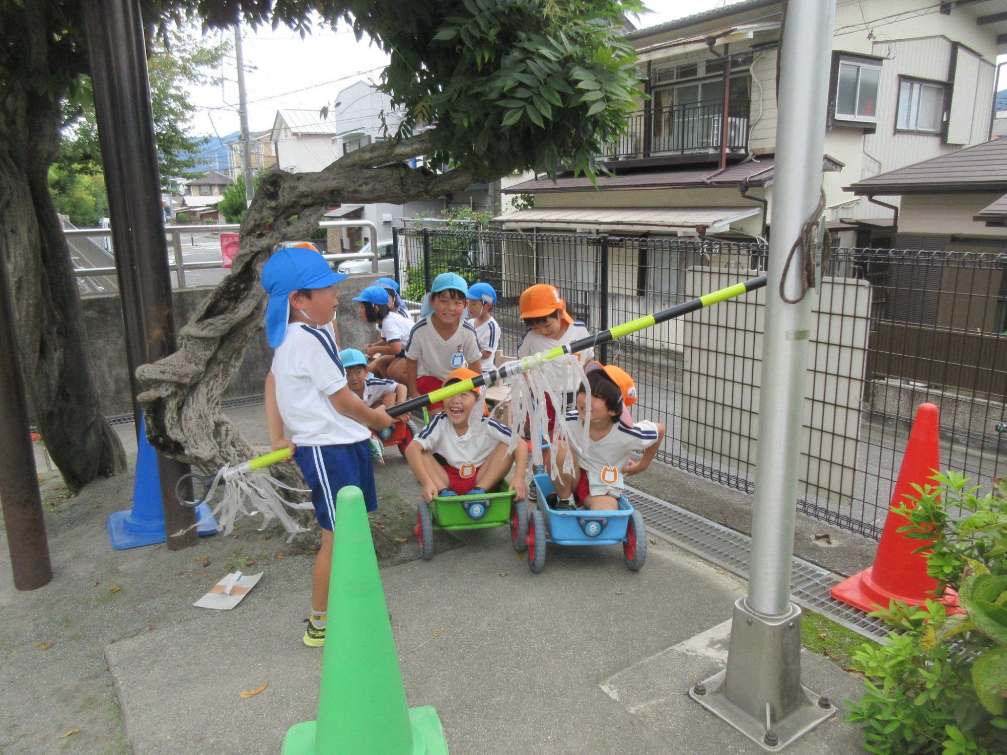 複数の園児が緑や水色の手押し車に乗り、その前で黄色と黒色のカラーコーンバーを持った園児が立ち、踏切に見立てて遊んでいる様子の写真