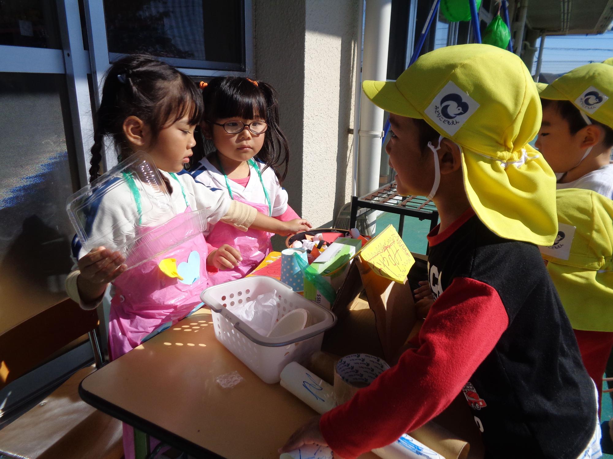 テラスのテーブルでピンクのエプロンを着た園児2人が、他の園児たちに紙コップや画用紙を使って作ったお寿司を見せている写真
