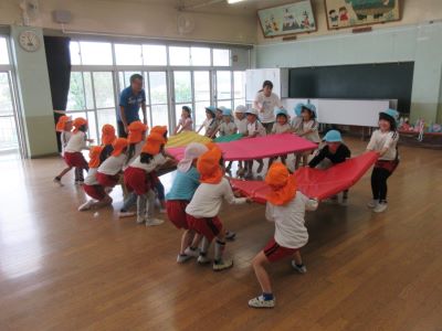 室内で複数の園児たちが5、6人ずつでカラフルな大きなマットを持ち、引っ張り合って遊んでいる写真